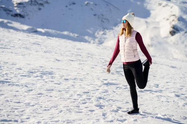 Full body of young female traveler in cozy clothes and beanie cap, standing on one leg and holding leg up while looking away on snowy slope in daylight