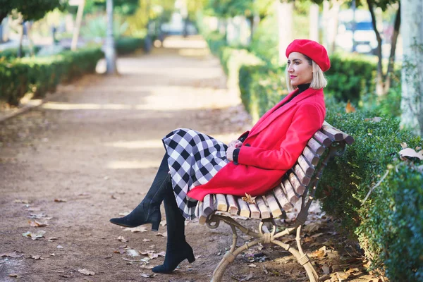 緑の植物と秋の公園の木のベンチに座っている赤い衣装とベレーの女性のフルボディサイドビュー — ストック写真