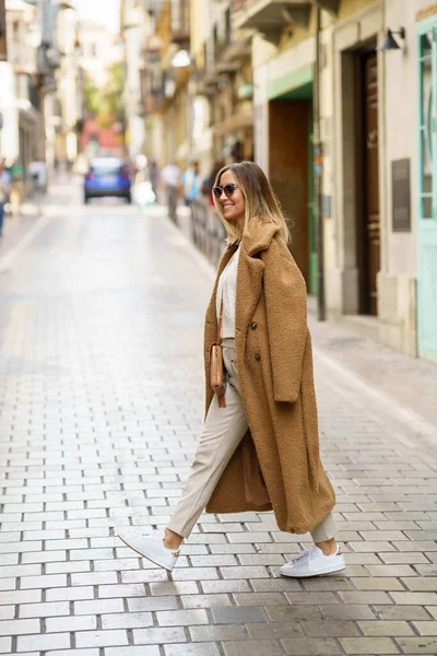 Moda montlu, güneş gözlüklü ve omuz çantalı genç bir kadın caddede karşıdan karşıya geçerken görülüyor.