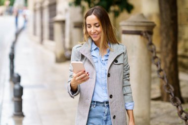 Mutlu, yetişkin bir kadın, günlük kıyafetler ve şık gri bir ceketle, şehrin bulanık arka planında dikilirken akıllı telefondan gülümsüyor ve video seyrediyor.