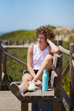 Beyaz bluzlu, kıvırcık saçlı ve siyah şortlu, su şişesinin yanındaki ahşap bankta oturan, bulanık arkaplana bakan, pozitif genç bir kadın.