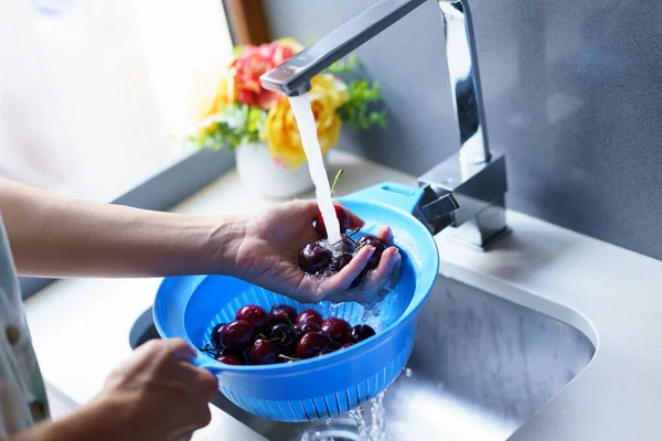 From above of crop anonymous woman washing ripe cherries in plastic colander under water from faucet in kitchen sink