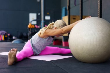 Spor giyimli çıplak ayaklı bayan sporcunun yan görünüşü, bacaklarıyla mindere uzanmış, elleri topun üzerinde otururken ve gün ışığında spor salonunda esneme egzersizi yaparken.