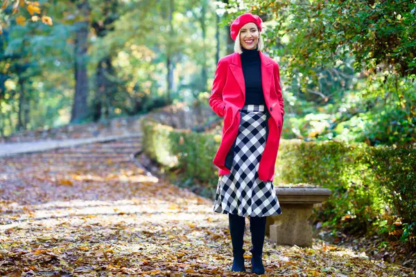 秋の公園の落ち葉の上に立っているチェッカー付きのスカートで赤いコートとベレーを身に着け 幸せに微笑むポジティブな女性の全身 — ストック写真