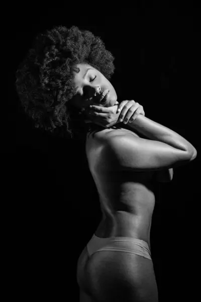 Topless black woman with Afro hairstyle closing eyes under studio light against black background. Black and white photograph.