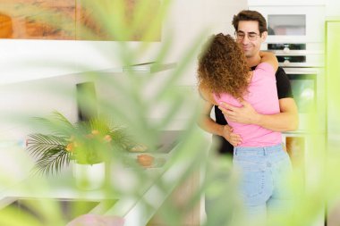 Modern apartmanın terasında yeşil bitkiler ve çiçeklerin yanında dikilen, günlük giysiler içinde gülümseyen genç çiftin manzarasına bakın.