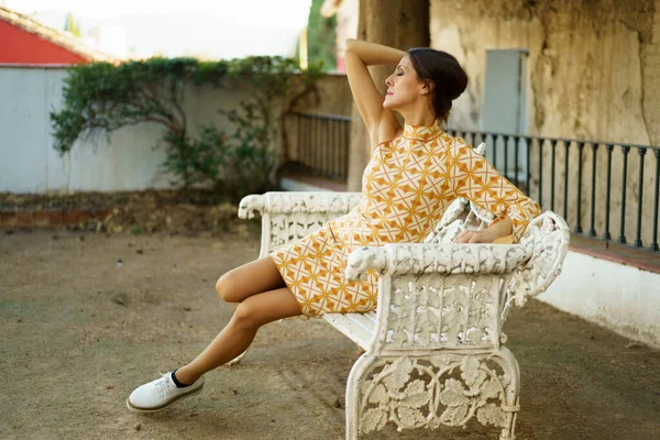 Full body of young alluring female model in stylish dress and sneakers sitting up in romantic pose with closed eyes on bench by old building