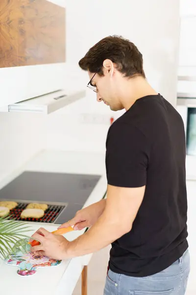 カジュアルな服と眼鏡で見下ろす大人の男性の側面 調理範囲の近くのキッチンに立っている間 グリルにパンをスライスし 昼光でナイフでトマトを切る — ストック写真