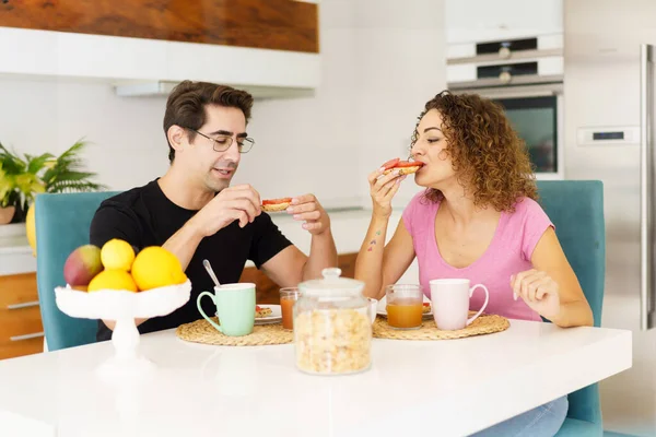 Happy adult couple in conversation and looking down while sitting at dining table in kitchen with juice in glasses, on placemat near fruits in bowl and eating salad in daylight at home