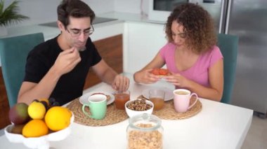 Gerçek zamanlı mutlu yetişkin çift, yemek masasında otururken birbirlerine bakıyorlar kahve bardakta kahve bardakta kahve ve gün ışığında meyve reçeliyle yayılmış ekmek dilimleri yiyorlar.