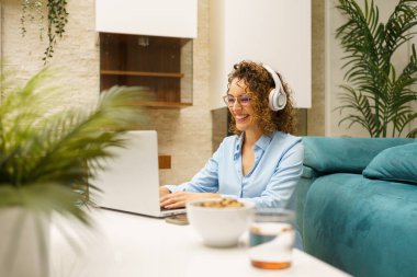 Neşeli kıvırcık saçlı, günlük kıyafetleri ve kulaklıkları olan serbest çalışan, modern, rahat oturma odasında dizüstü bilgisayarlı, uzaktan kumandalı bir proje üzerinde çalışan bir kadın.