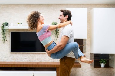 Modern dairede LED TV karşısında romantik bir an yaşarken kucaklaşan gülümseyen genç çiftin yan görüntüsü ve birbirlerine mutlu bir şekilde bakmaları.