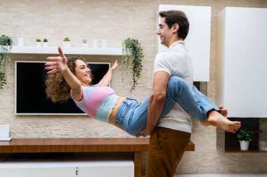 Olumlu genç çiftin birlikte eğlenip eğlenirken evlerinde saksı yeşili bitkilerle duvarda kurşun televizyonun karşısında dikilirken yan görüntüsü.