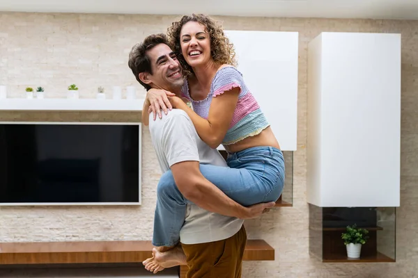 カジュアルな服で若い陽気なカップルが抱擁し 自宅に立って 棚に植物を置いたテレビと一緒に時間を楽しんでいる側面 — ストック写真