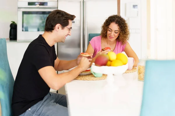 Happy adult couple in conversation and looking down while sitting at dining table in kitchen with fruits in bowl, and eating breakfast in daylight at home