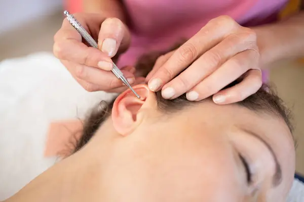 Crop professional chiropractor applying auriculotherapy ear acupuncture techniques on female client lying in modern beauty salon during skin care treatment