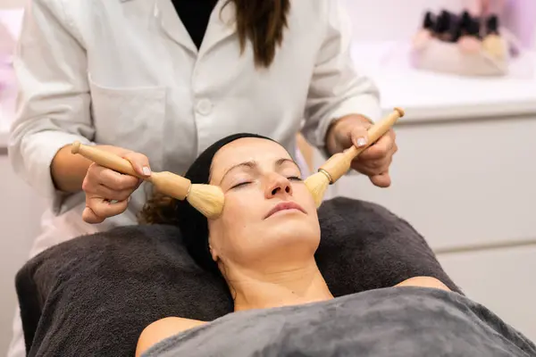 Crop anonymous cosmetician in uniform massaging face of relaxed woman with brushes during facial treatment at beauty salon