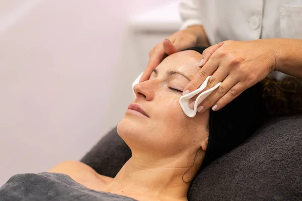 Crop anonymous cosmetician removing skin care product from face of female client with cotton pads during professional facial treatment