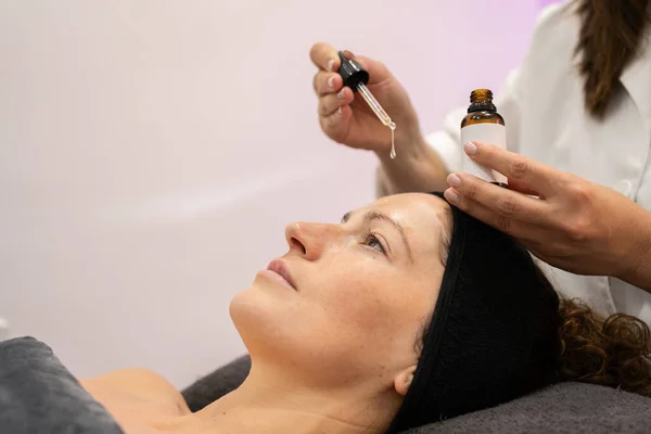 Crop beauty specialist applying serum on face of young female customer lying in modern salon during skin care treatment
