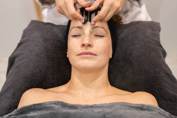 High angle of crop anonymous female cosmetician doing facial massage with black crystals during anti aging skin care procedure in beauty salon