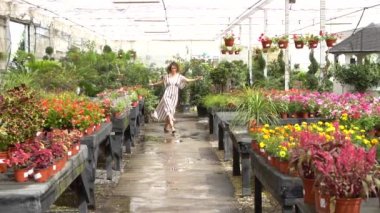 Yavaşça gülümseyen genç kadın, el kamerasıyla kıvırcık saçlara dokunurken ve el hareketleriyle gün ışığında bulanık çiçeklere karşı veda ederken.