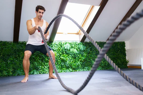 Full body of young fit male athlete in activewear doing exercise with battle ropes during intense training in gym with pitched roof