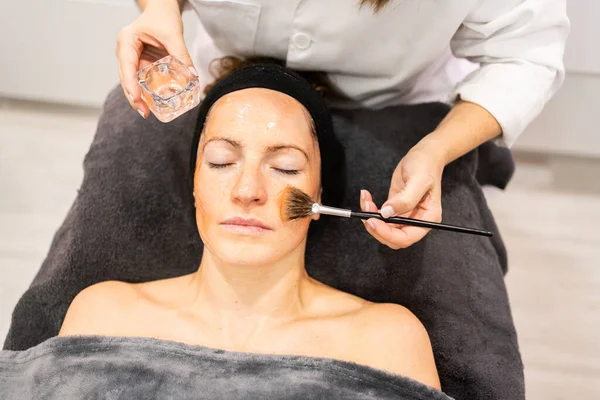 From above anonymous beautician applying liquid cosmetic gel with brush on woman face during skin care treatment in beauty salon