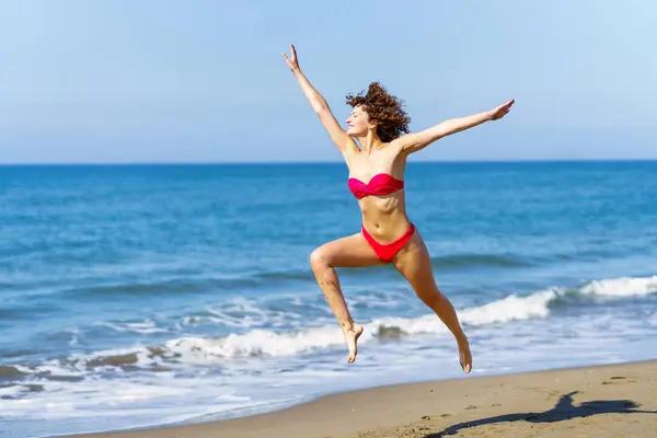 笑顔のケアフリー ピンクのビキニで若いスリム女性の赤毛 腕を上げて 離れて見ている間に空気中の足でジャンプ ぼやけた海と青空に対して日光で — ストック写真