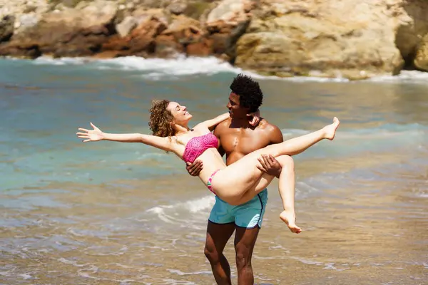 腕を後ろに伸ばす陽気な若い女性の側面は アフリカ系アメリカ人の男性の手中に横たわり 昼間の海水の近くで互いを見ています ストック写真