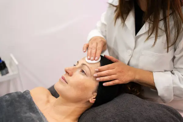 スキンケア治療中に美容院で顔の洗浄手順のための女性クライアントの顔にコットンパッドを使用して作物宇宙医 ストックフォト