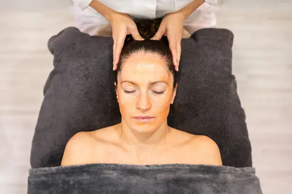 上記から クロップビューティスペシャリストマッサージヘッド タオルに横たわる女性クライアント スキンケア治療中に美容センターで目を閉じた ストック画像