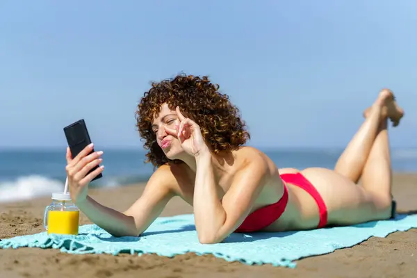 海岸に横たわるビキニの若いカーリーヘアの女性と日光浴中にセルフィーを取っている間に2本の指を示す ロイヤリティフリーのストック写真