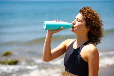 Dalgalı denizin yanındaki eğitimden sonra plastik şişeden su içen siyah tişörtlü, kıvırcık saçlı genç bayan sporcunun yan görüntüsü.