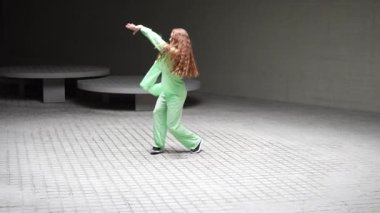 Uzun kızıl saçlı bir kadın modern, minimalist bir ortamda dans gösterisi yapıyor. Parlak yeşil kıyafeti nötr arkaplan karşısında çarpıcı bir görsel etki yaratıyor..