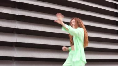 Canlı yeşil kıyafetli bir kadın, yatay çizgili modern bir binanın önünde enerjik bir şekilde dans eder. Yaşam tarzı, moda ve kentsel sahne içeriği için mükemmel.