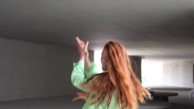 Şehir tünelinde, parlak kırmızı saçlı genç bir kadın enerjik bir şekilde yeşil bir ceket ve siyah bir bluzla dans ediyor, neşe ve hareketlerle dolu, tasasız ve spontane bir sahne canlandırıyor.