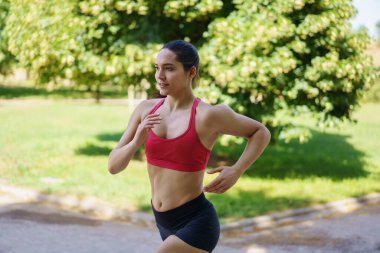 Kırmızı spor sutyen ve siyah şortlu genç bir kadın güneşli bir günde yeşil bir parkta koşuyor, spor ve sağlıklı bir yaşam tarzını destekliyor, aktif, egzersiz, sağlık ve sağlık gibi anahtar kelimeleri somutlaştırıyor.