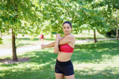 Yemyeşil bir parkta, kırmızı spor sutyen ve siyah şortlu genç bir atletik kadın kolunu uzatıyor, açık hava antrenmanına hazırlanıyor, spor ve kuvvete olan bağlılığını gösteriyor.