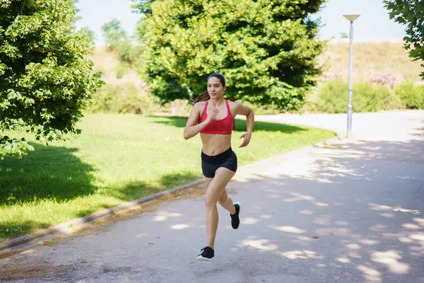 Spor giyimli genç bir kadın güneşli bir parkta kararlı bir şekilde koşuyor. Forma ve sağlıklı bir yaşam tarzına olan bağlılığını gösteriyor. Sağlığına olan bağlılığı, enerjik adımlarında belli oluyor.