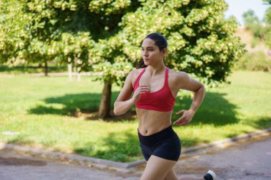 Aktif bir genç kadın parkta koşuyor, vücut bulmuş spor yapıyor ve sağlıklı bir yaşam sürüyor. Kırmızı spor sutyen ve siyah şort giymişti. Egzersizine odaklanmış ve kararlı görünüyordu.