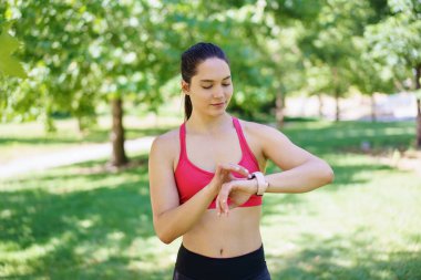 Canlı yeşil bir parkta, kendini pembe spor sutyenli genç bir kadın spor takip cihazını kontrol ederken sağlık ve zindelik rutinine odaklanıyor. Açık havada aktif kalmaya kendini adamış.