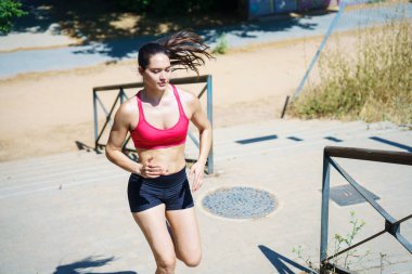 Spor giyim sektöründe kararlı bir bayan sporcu spor salonuna odaklanmış, parkta merdivenlerden yukarı koşuyor. Kendini açık hava egzersizine adadı. Güneşin altında gücünü ve sağlığını koruyor.