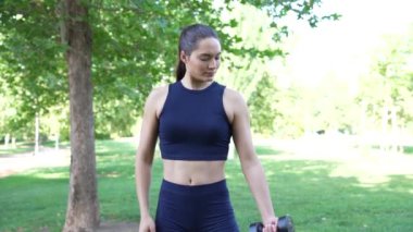 Formda genç bir kadın, yeşil bir parkta halterlerle biseps kıvrımları sergiliyor. Açık hava sporunu gösteriyor.