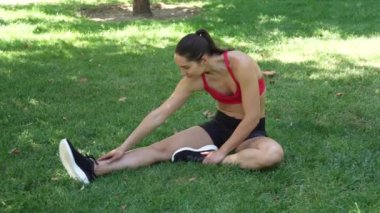 Bir kadın, bir parktaki çimenli bir alanda spor yapıyor, fitness, esneklik ve açık hava sağlığını teşvik ediyor. Görüntüler onu kırmızı atlet ve siyah şortla gösteriyor..