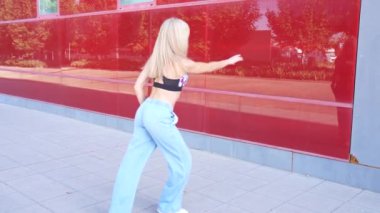 Gündelik giysiler içinde enerjik genç bir kadın modern bir binanın yanında kendinden emin bir şekilde dans ediyor. Canlı bir şehir ortamında dinamik hareketleriyle özgürlüğünü ve neşesini ifade ediyor.
