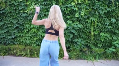 Uzun sarı saçlı genç bir kadın gür, yeşil sarmaşık kaplı bir duvarın önünde enerjik bir şekilde dans ediyor. Şık, geniş bacaklı kot pantolon ve renkli bir bluz giyiyor. Açık hava manzarasının tadını çıkarıyor..