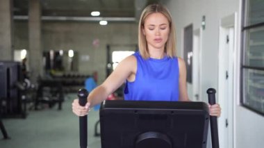Mavi spor kıyafetli aktif bir kadın modern bir spor salonunda eliptik bir makine kullanarak sağlığını korumaya odaklanır. Forma ve genel olarak sağlığa olan bağlılığını gösterir.