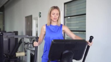Mavi askılı beyaz bir kadın modern bir spor salonunda eliptik bir makine üzerinde egzersiz yaparken odaklanmış ve kararlı bir şekilde egzersiz yaparak sağlıklı bir yaşam tarzına öncelik veriyor.
