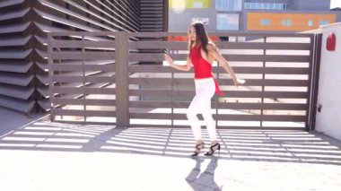 Parlak kırmızı bluzlu genç bir kadın çarpıcı bir modern mimari özelliğin yakınında jestler yaparak kendine has tarzını ve güvenini canlı ve enerjik bir açık hava ortamında sergiliyor.