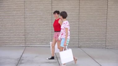 İki şık genç kadın, ellerinde son bulgularıyla dolu renkli alışveriş çantaları ve şık yaşam tarzlarını ve dostluklarını somutlaştıran moda kıyafetlerini taşıyarak işlek şehir sokaklarında dolaşıyorlar.